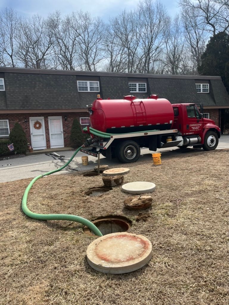 A red septic tank truck pumping a septic system on a grassy lawn in front of an apartment building.
