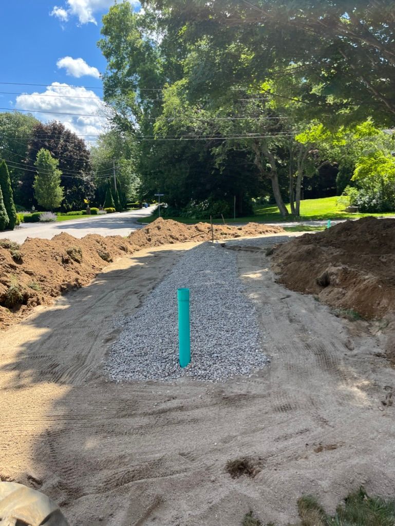 A gravel and sand pathway with a vertical green pipe, surrounded by dirt and green trees.