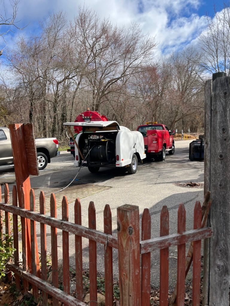 A white camper trailer with its door open, hitched to a red pickup truck, in a parking area with a wooden fence.