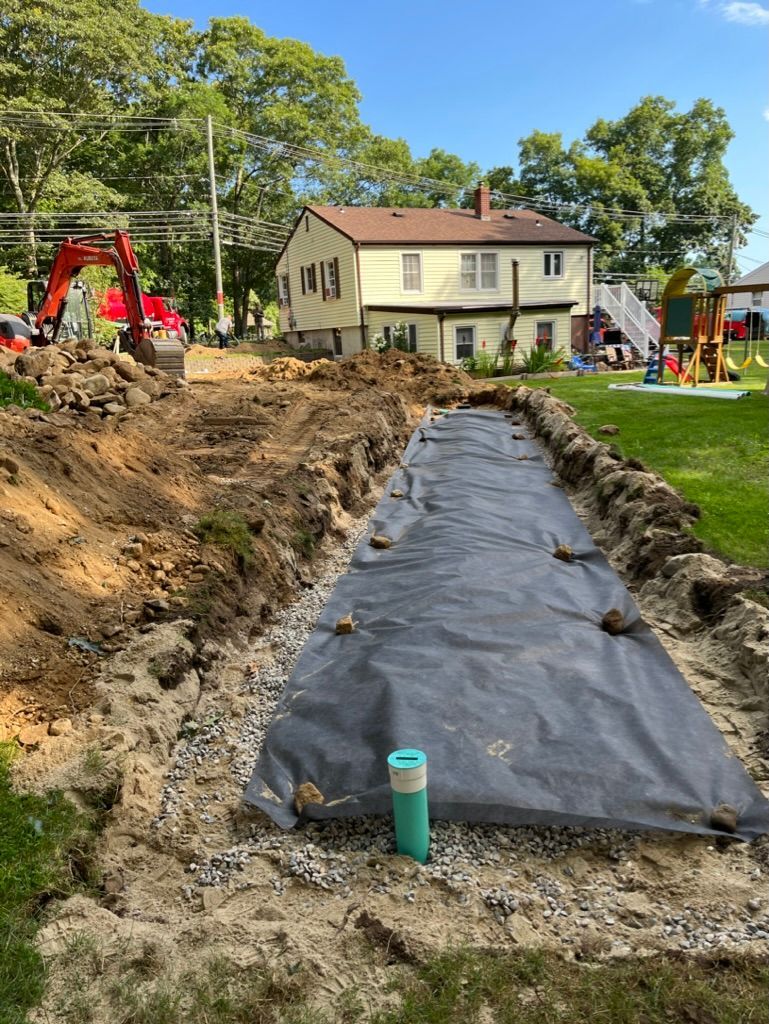 Construction site with trench covered in black fabric; backhoe, yellow house, and playground in the background.