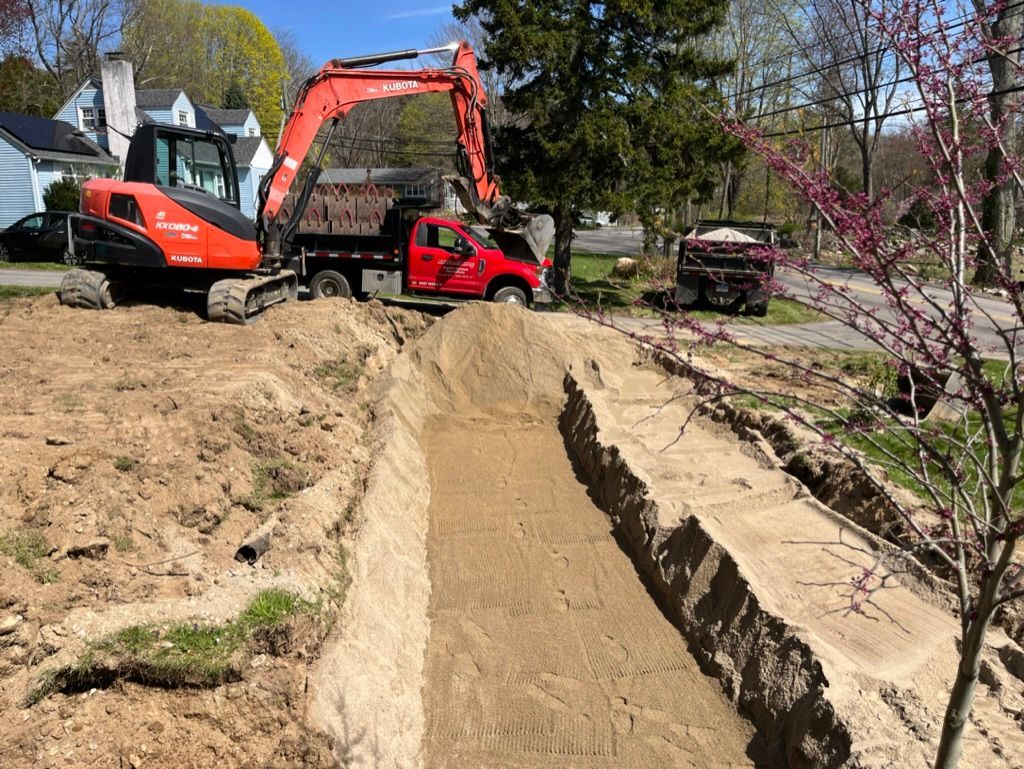 Orange excavator digging a trench, loading red dump truck with dirt in a residential setting on a sunny day.