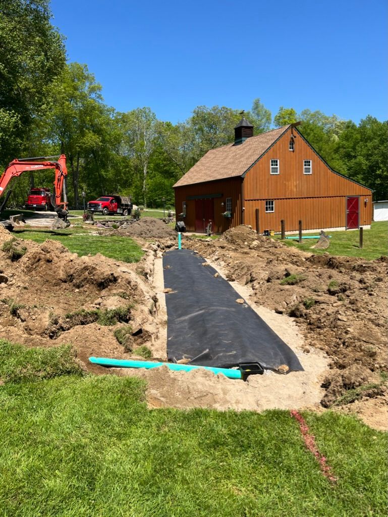 Construction site with trench, black liner, and green pipes; brown building in the background.