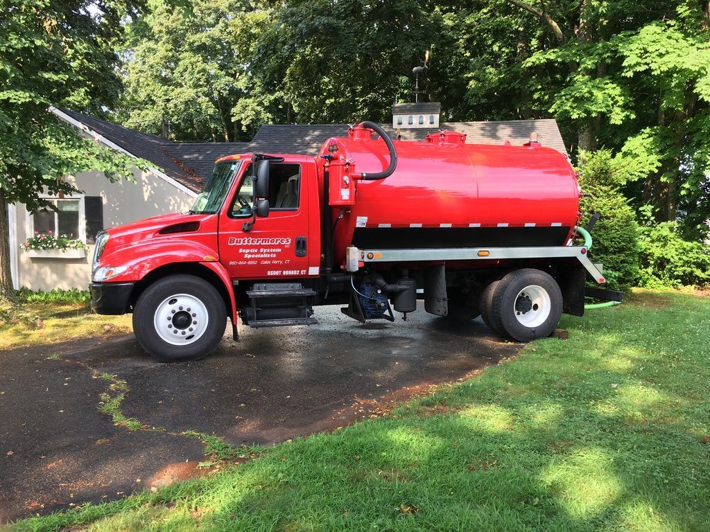Red septic tank truck parked in a residential driveway, next to a house and grassy lawn.