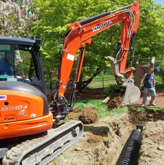 Orange Kubota excavator digging a trench, worker observing. Outdoor setting, trees in the background.