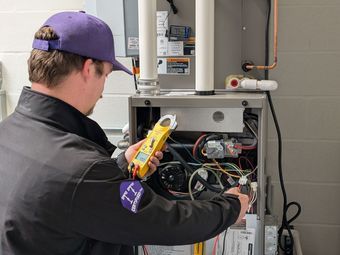 HVAC technician inspecting furnace with a multimeter, indoors. Wearing a purple hat, black jacket.