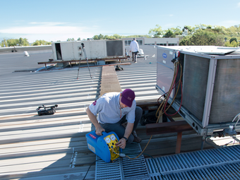 HVAC technician on a rooftop repairs a unit with blue equipment, sunny day.