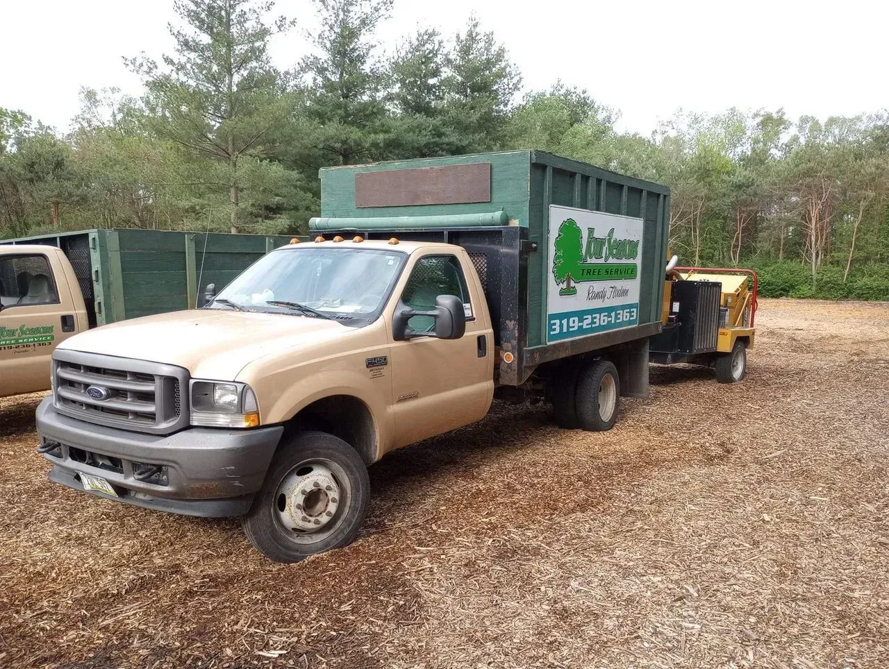 Tan truck with green dumpster bed and wood chipper trailer on a wood-chipped surface.