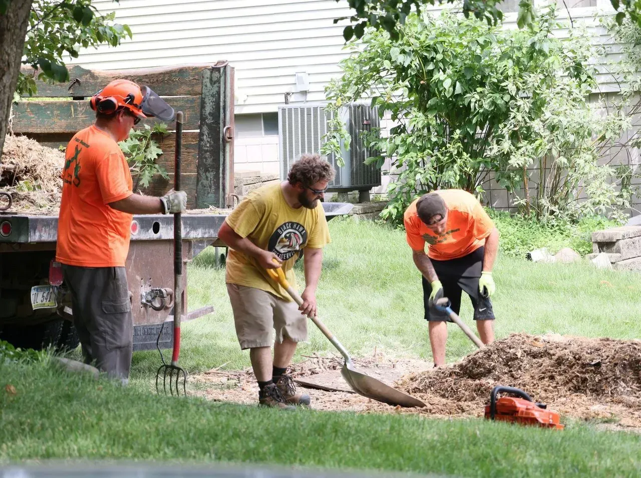 Three people spreading mulch with shovels near a truck and grassy area; one wears an orange shirt and helmet.