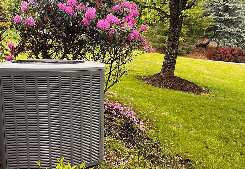 Outdoor air conditioner beside a green lawn and pink flowering shrubs in a landscaped yard