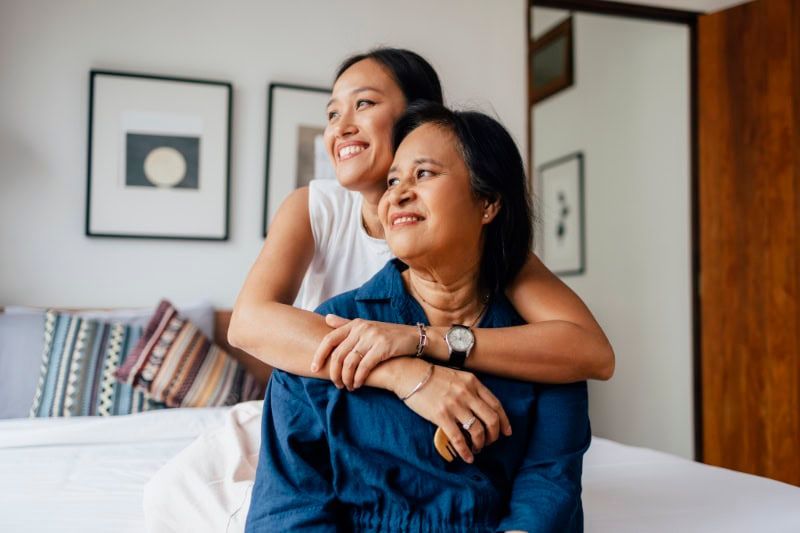 Two people sitting on a bed, smiling and hugging in a bright bedroom