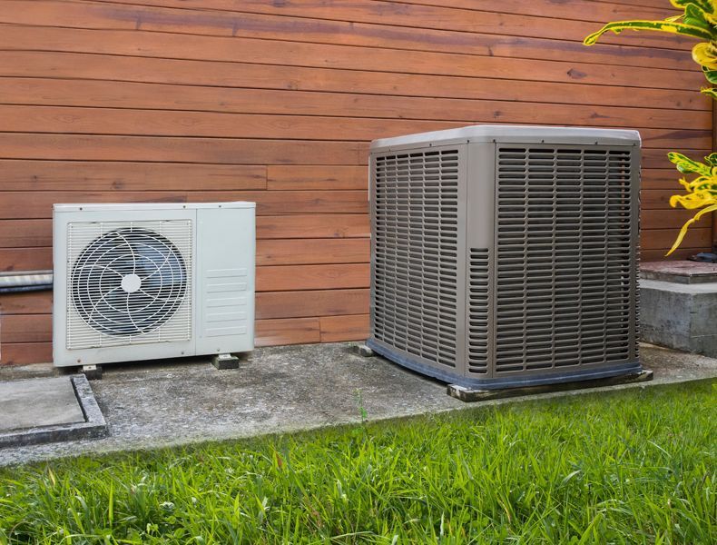 Two outdoor HVAC units beside a wooden wall, one white and one dark gray, on a concrete pad by grass.