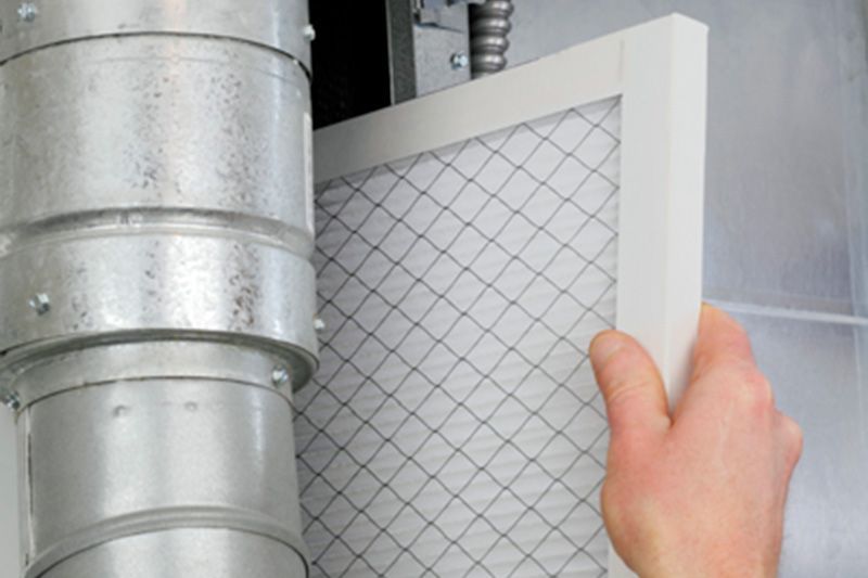 Hand holding a white air filter panel beside metal ductwork and a large HVAC unit