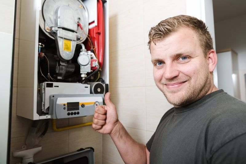 Man smiling beside an open furnace, giving a thumbs-up in a utility closet