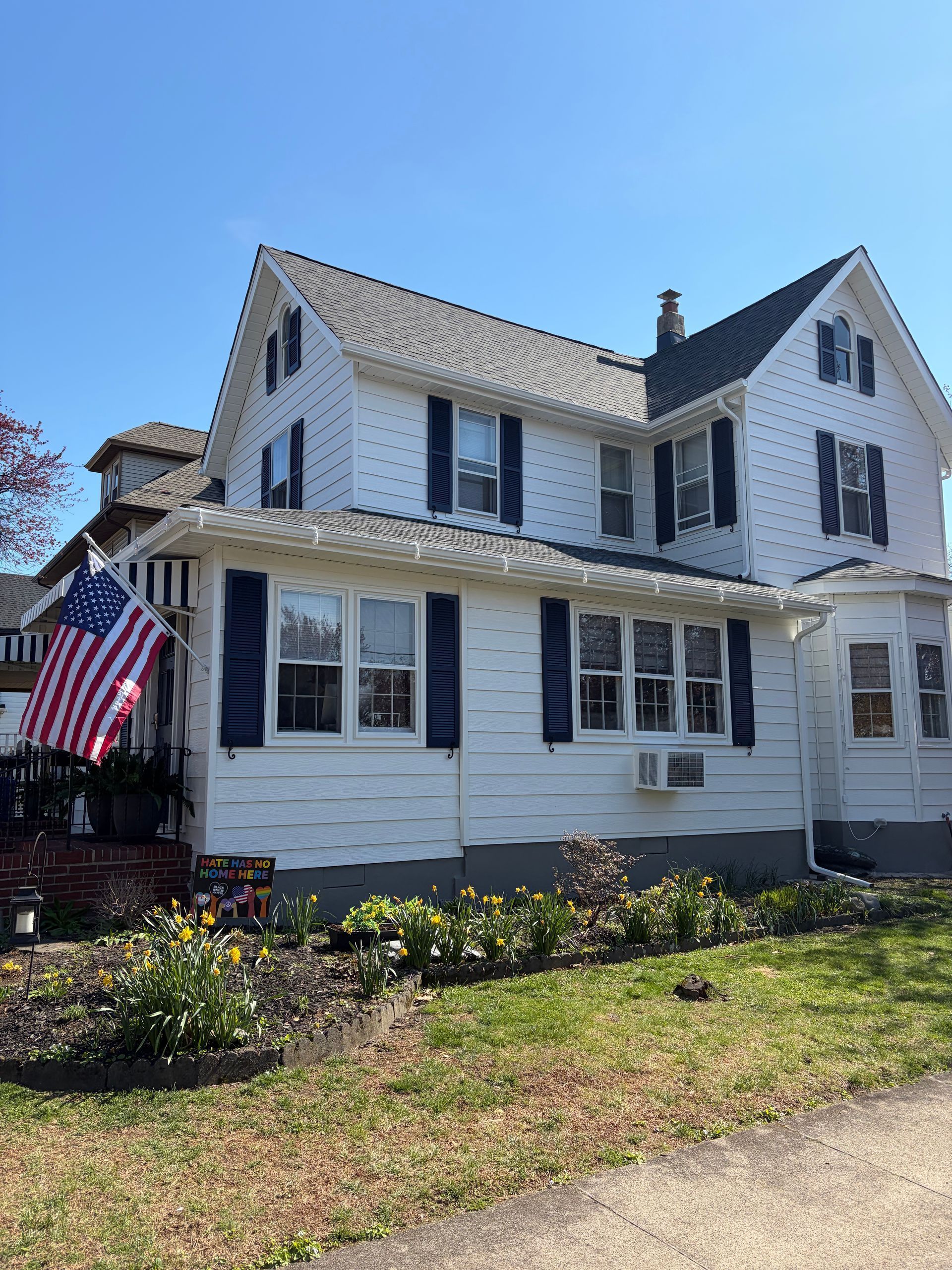 White two-story house with black shutters and a U.S. flag on the porch under a clear blue sky