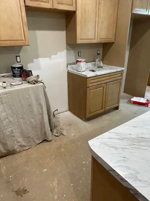 Kitchen under renovation; light brown cabinets, beige walls, marble countertops. Paint cans and drop cloths visible.