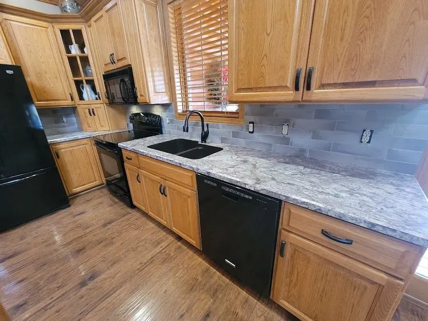 Kitchen with light wood cabinets, gray countertops and backsplash, black appliances, and wooden blinds.