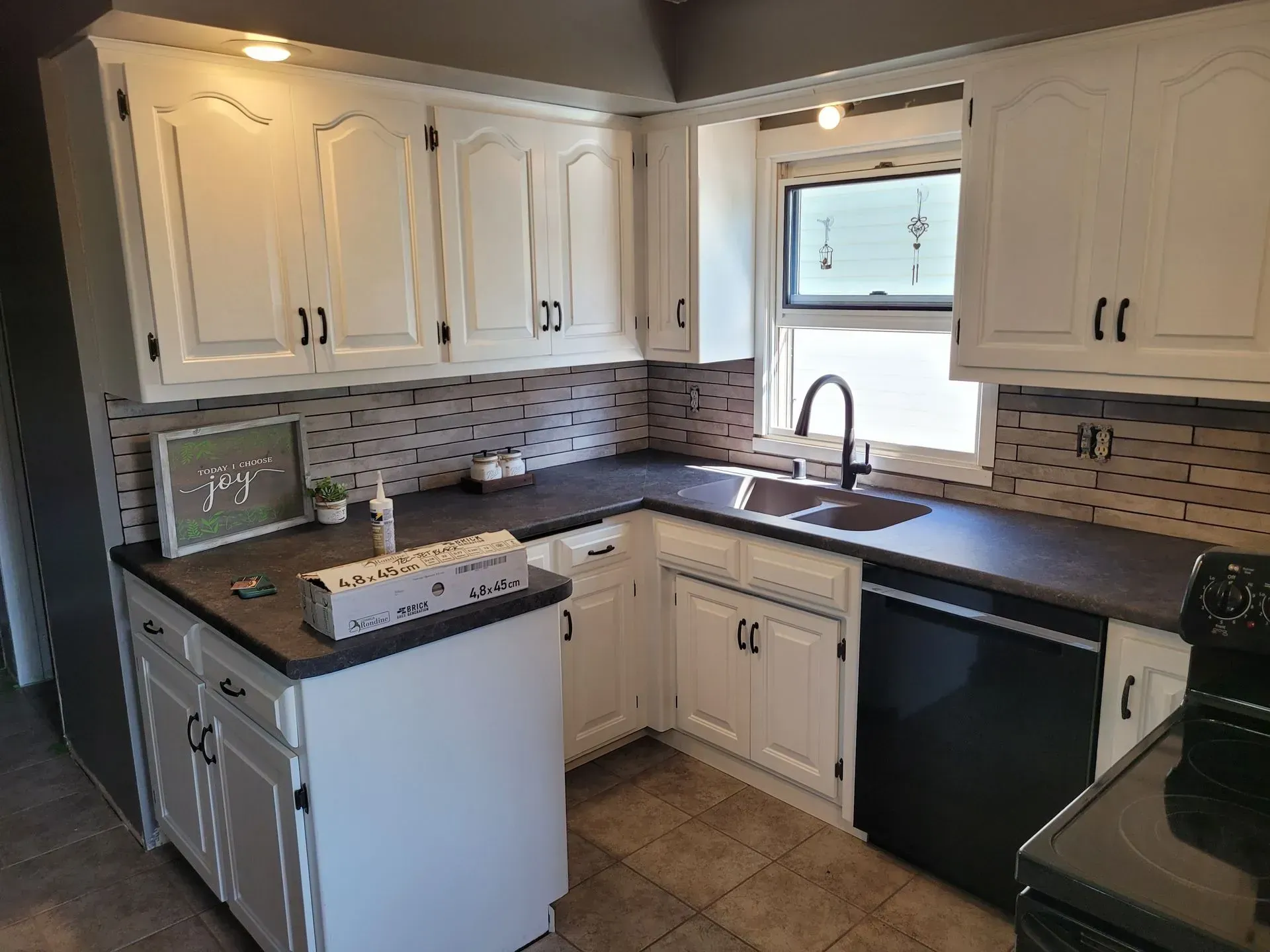 White kitchen cabinets with black hardware and appliances, dark countertops, brick-like backsplash, and a window.
