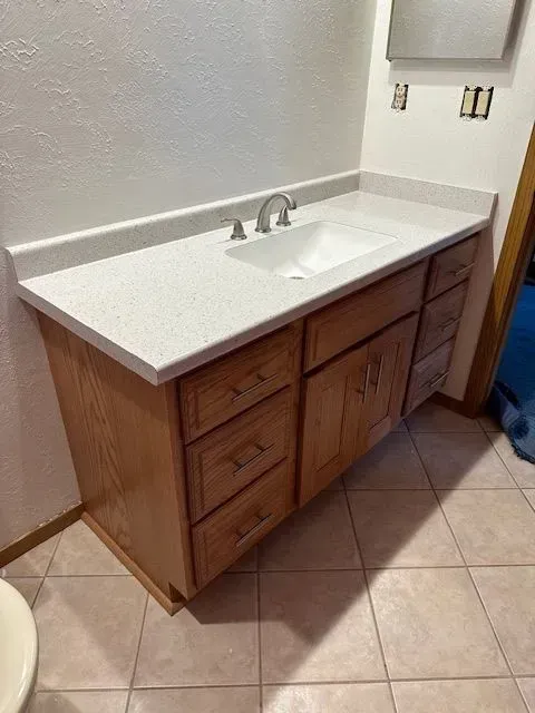 Wooden bathroom vanity with white countertop, sink, and faucet. Beige floor, white walls.