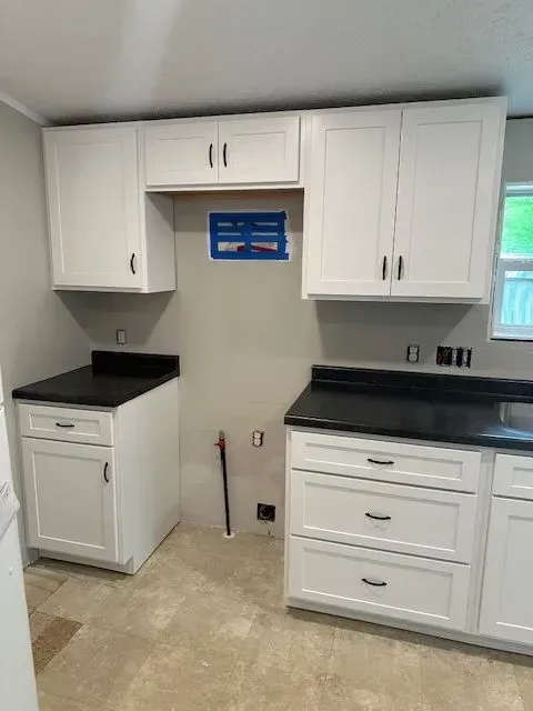 White kitchen cabinets with black countertops. Unfinished space, visible plumbing, and electrical outlets.
