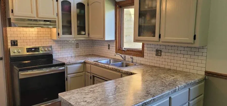 U-shaped kitchen with white cabinets, light-colored countertops, and subway tile backsplash. Stainless steel range and window.