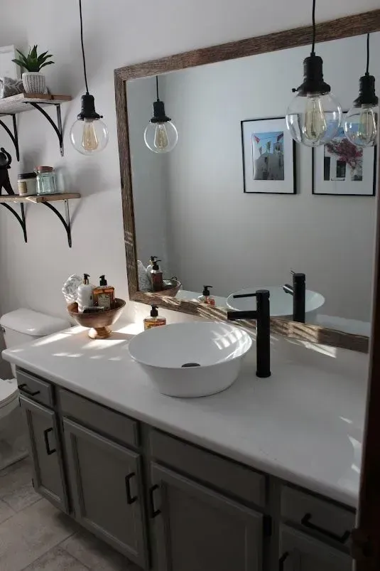 Bathroom with gray vanity, white countertop, and vessel sink. Large mirror, pendant lights, and floating shelves.
