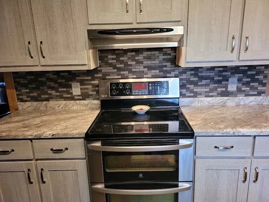 Kitchen with stainless steel double oven, stove, and grey tile backsplash. Light-colored cabinets and countertops.