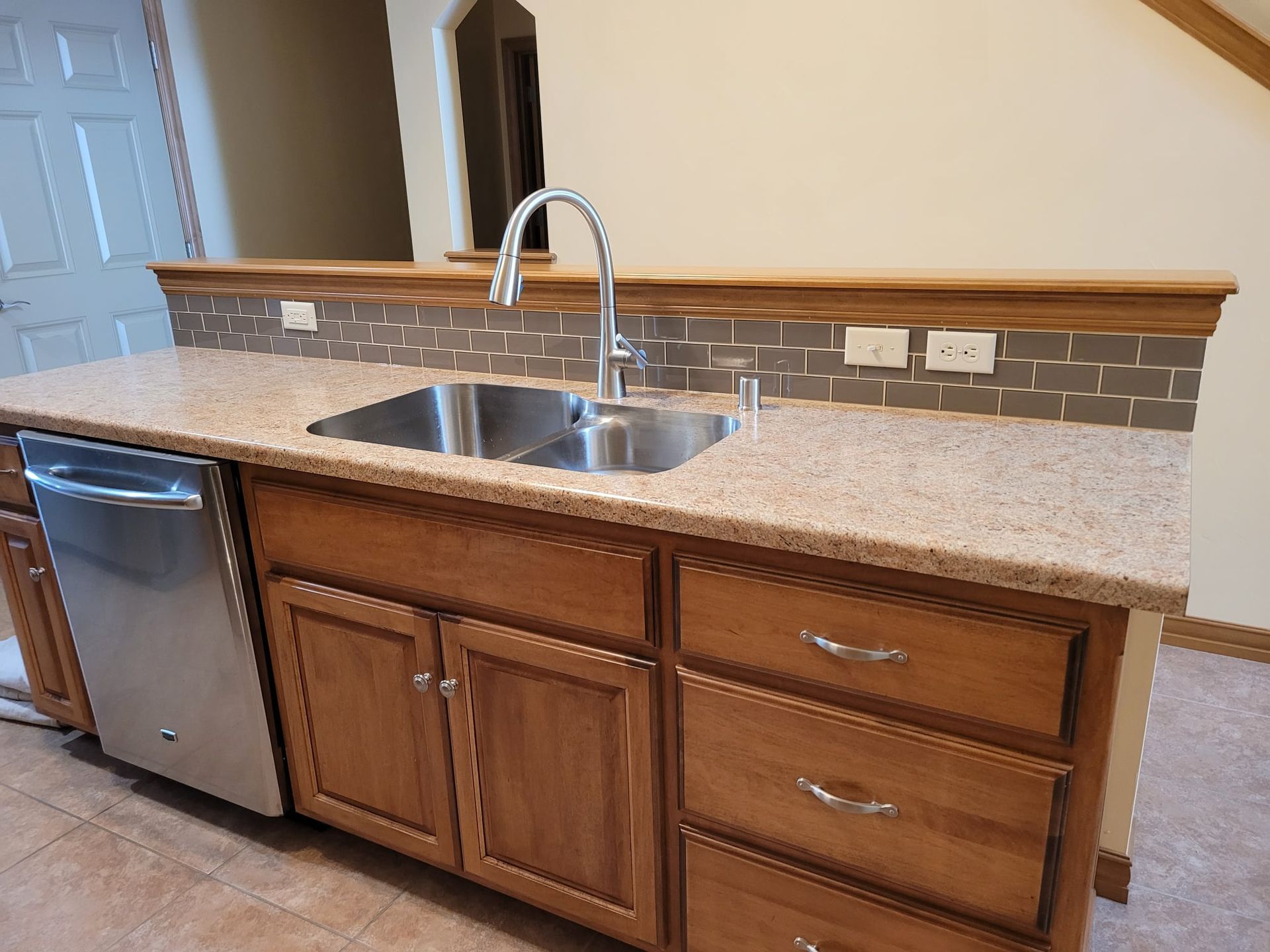 Kitchen with light-colored countertop, stainless steel sink, and wooden cabinets. Dishwasher on the left.
