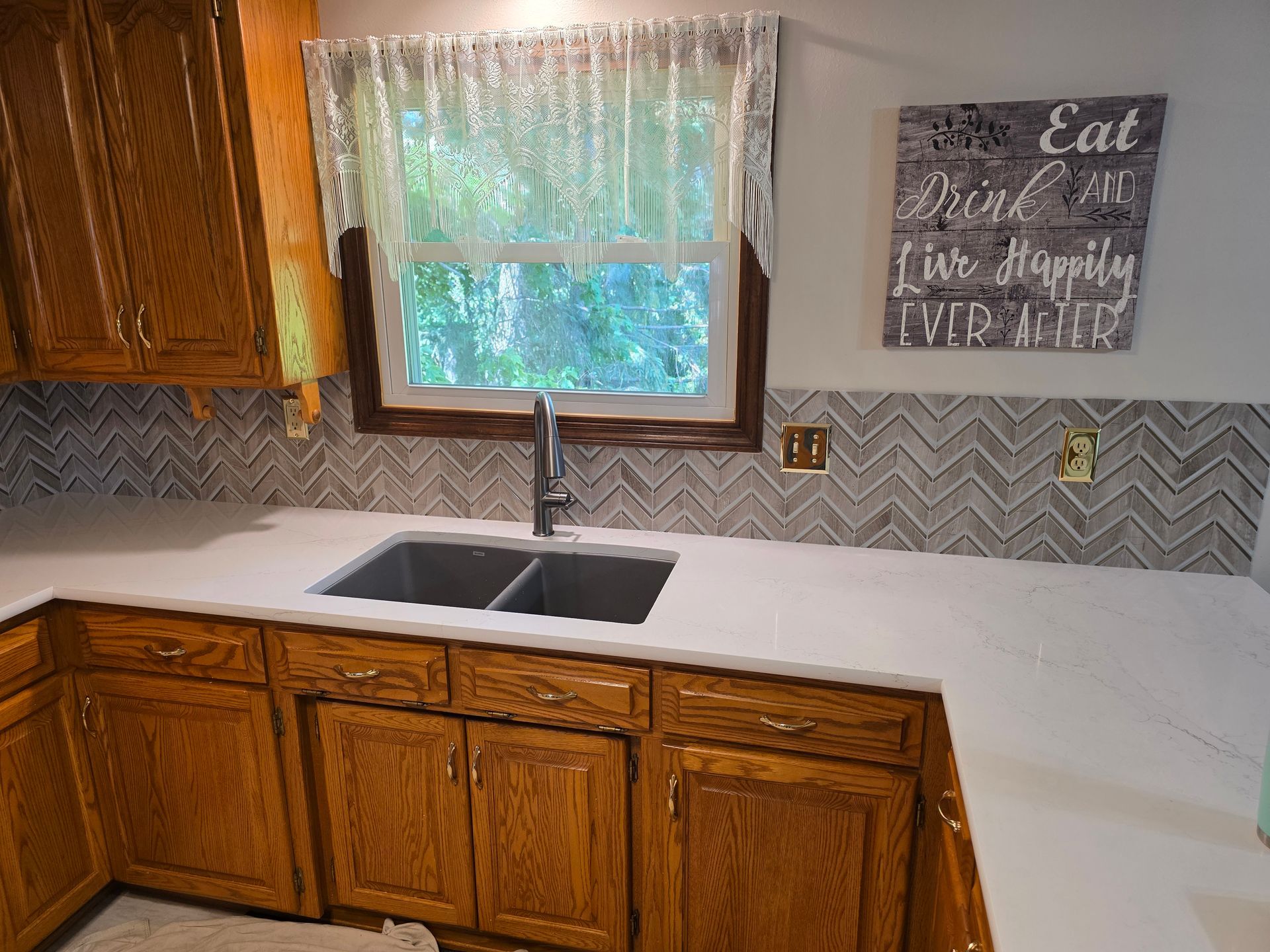 Kitchen with wooden cabinets, white countertop, sink, herringbone backsplash, and lace curtain.