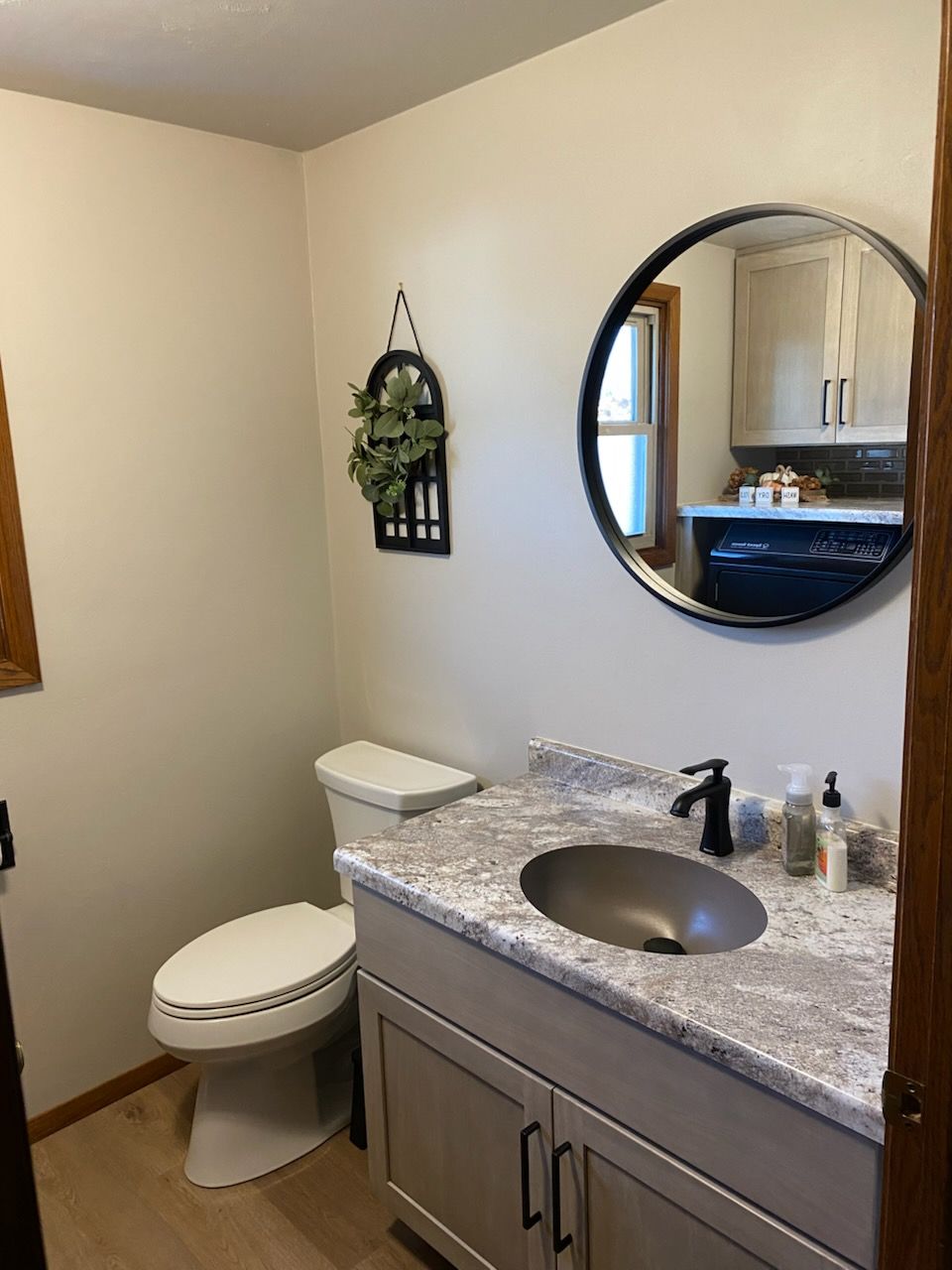 Bathroom with light-colored walls, toilet, vanity with round mirror, and decorative wall plant.