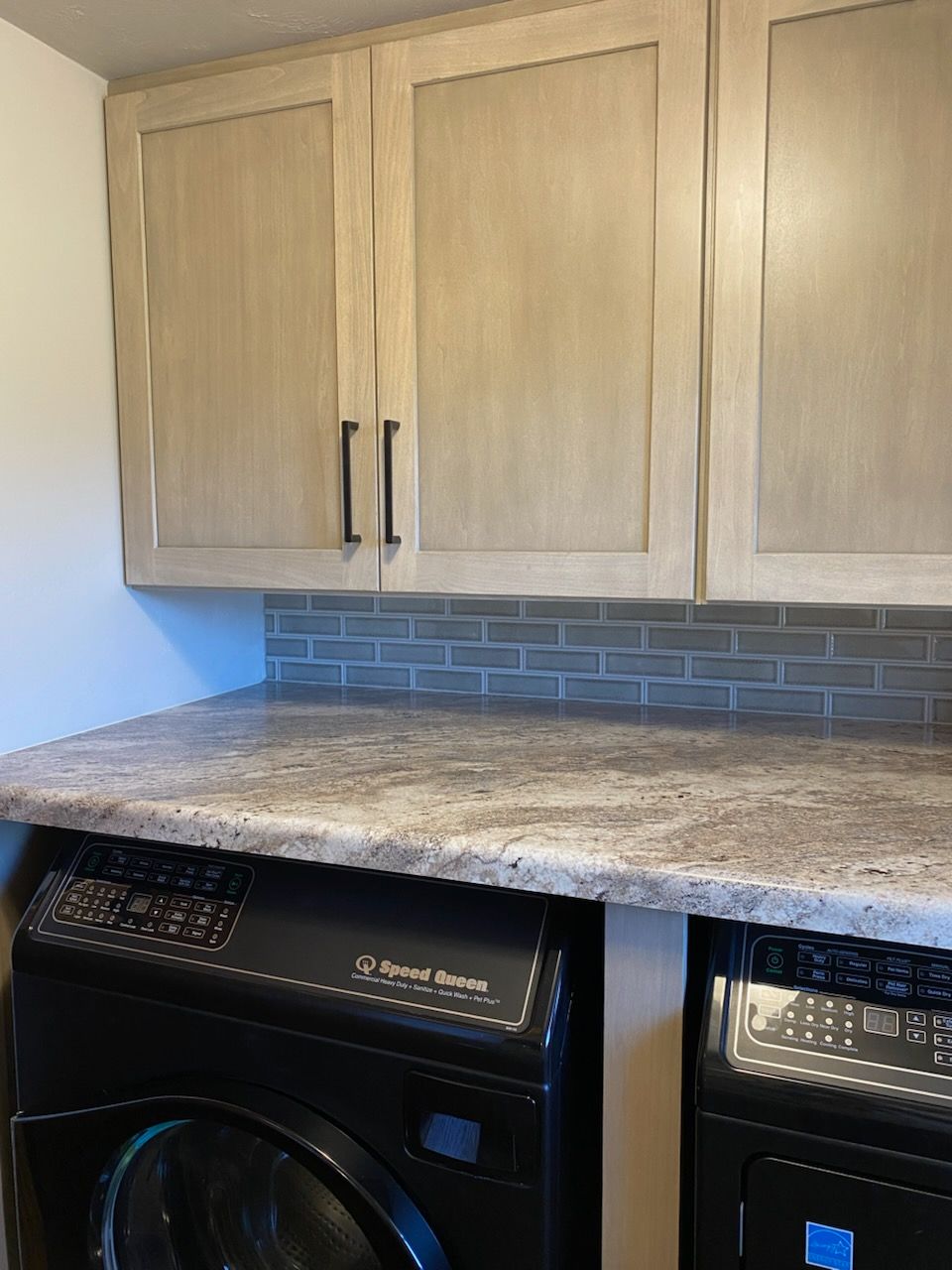 Laundry room with light wood cabinets, gray countertops, and black washer/dryer units.