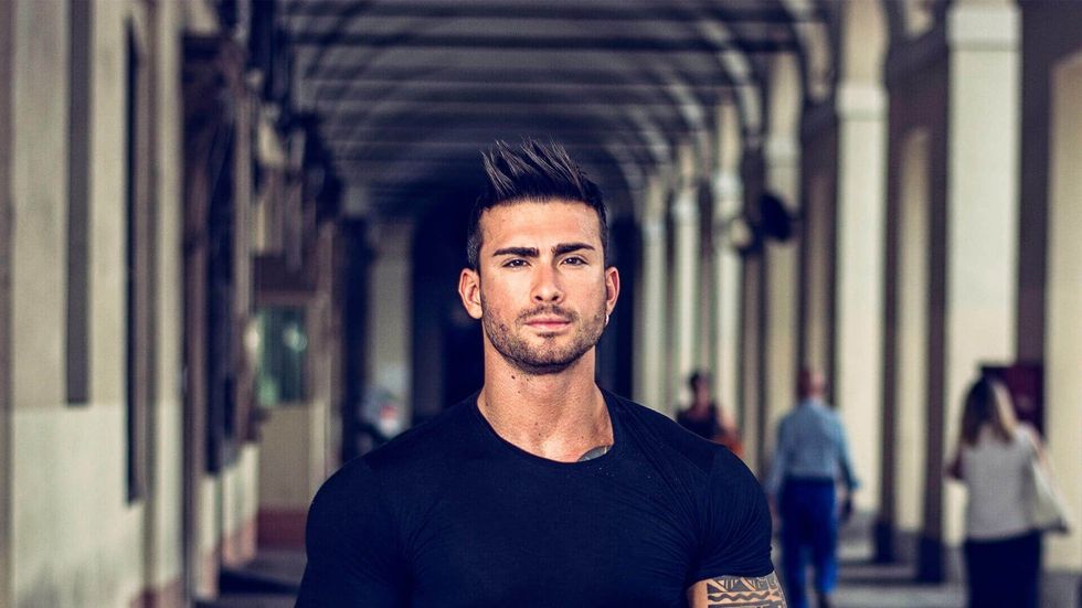 A muscular person with a short, spiked hairstyle stands in a sunlit, long stone hallway with blurred figures in the back.