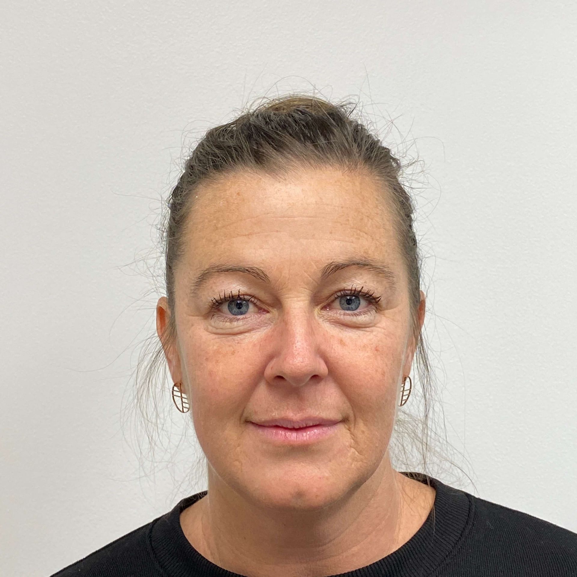 A person with tied-back brown hair and silver hoop earrings, looking directly at the camera against a plain white background.