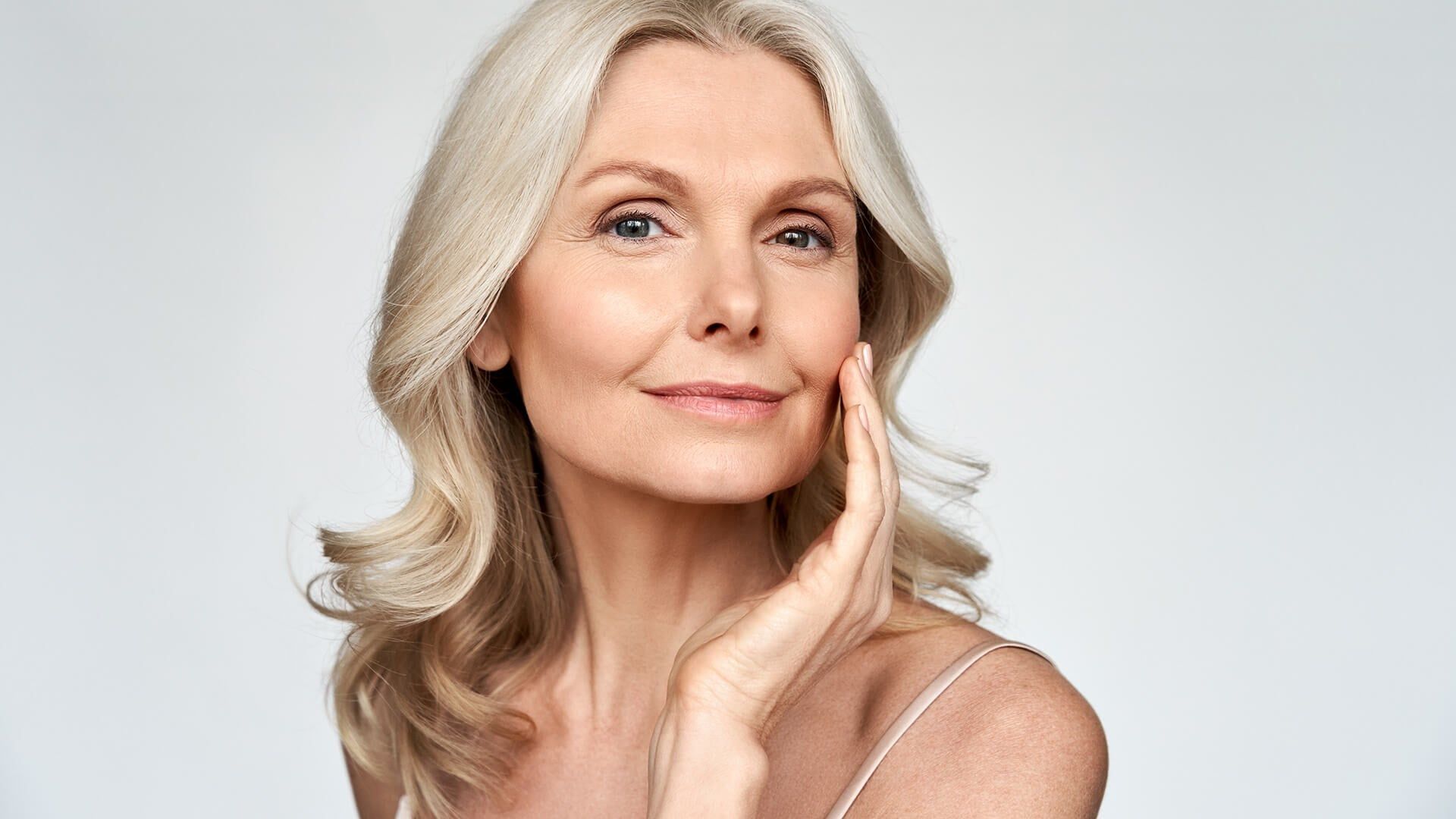 A woman with silver hair touching her cheek while looking at the camera against a light gray background.