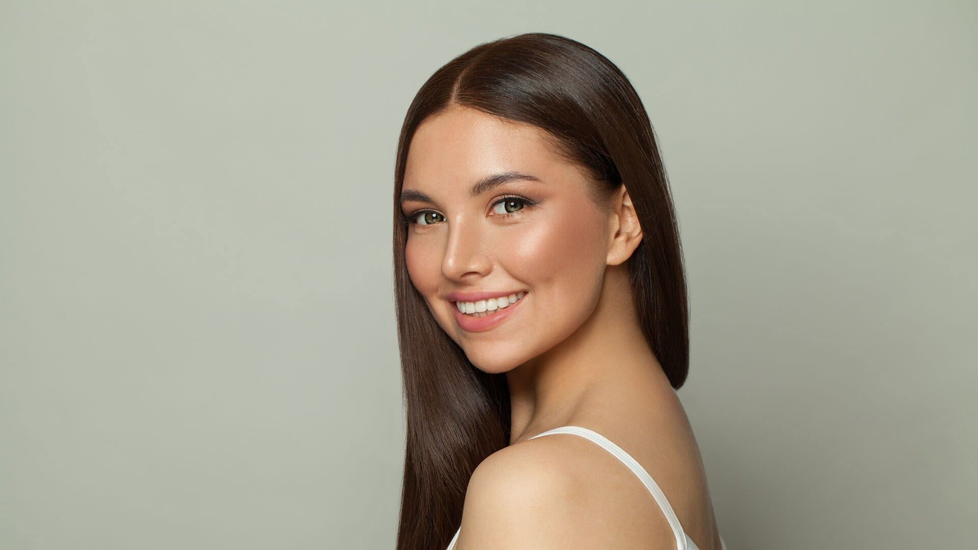 A smiling person with long dark hair, wearing a white top against a soft, light-green studio background.