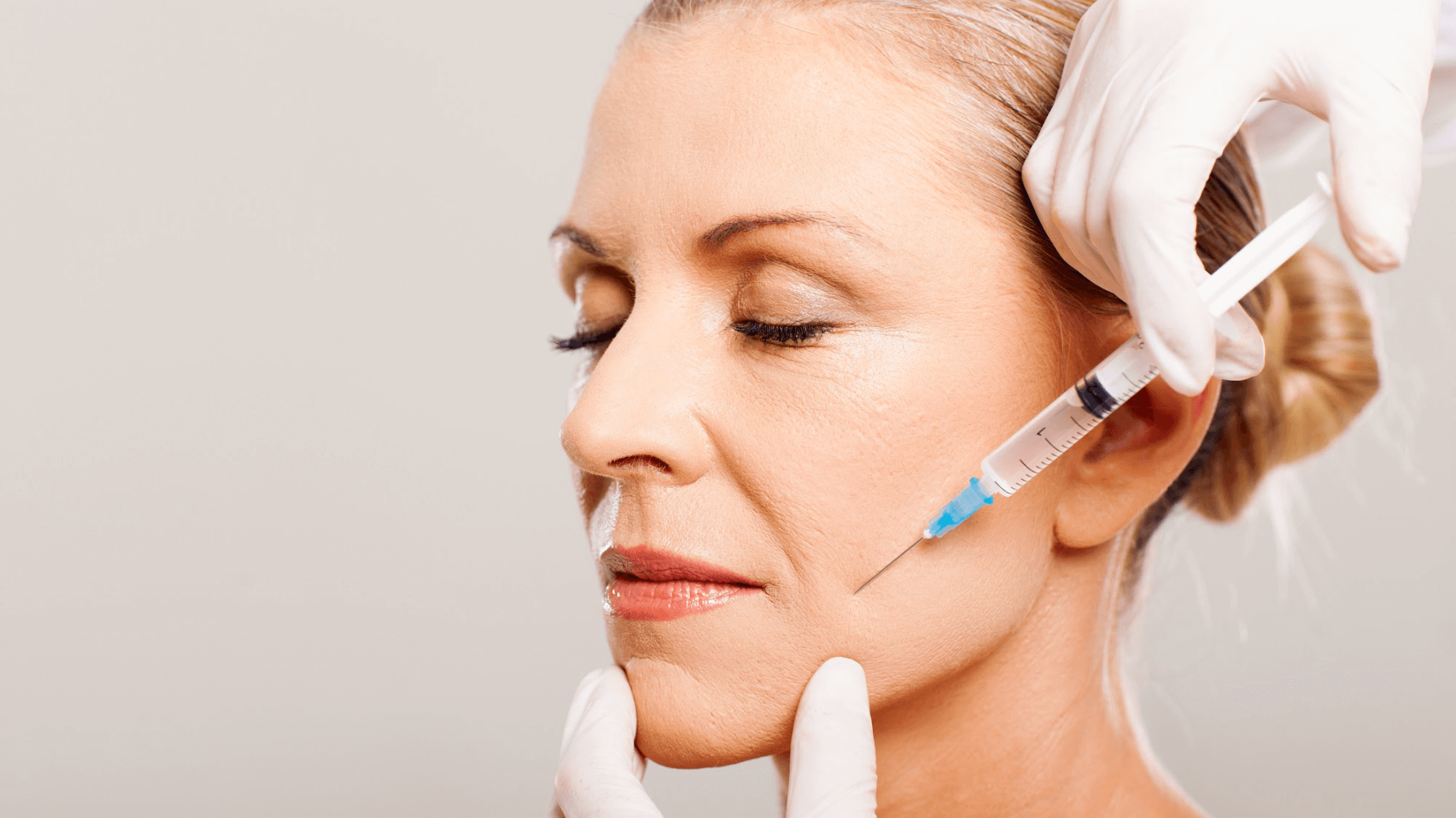 A clinician in gloves administers a cosmetic facial injection to a person near the cheek and jawline.