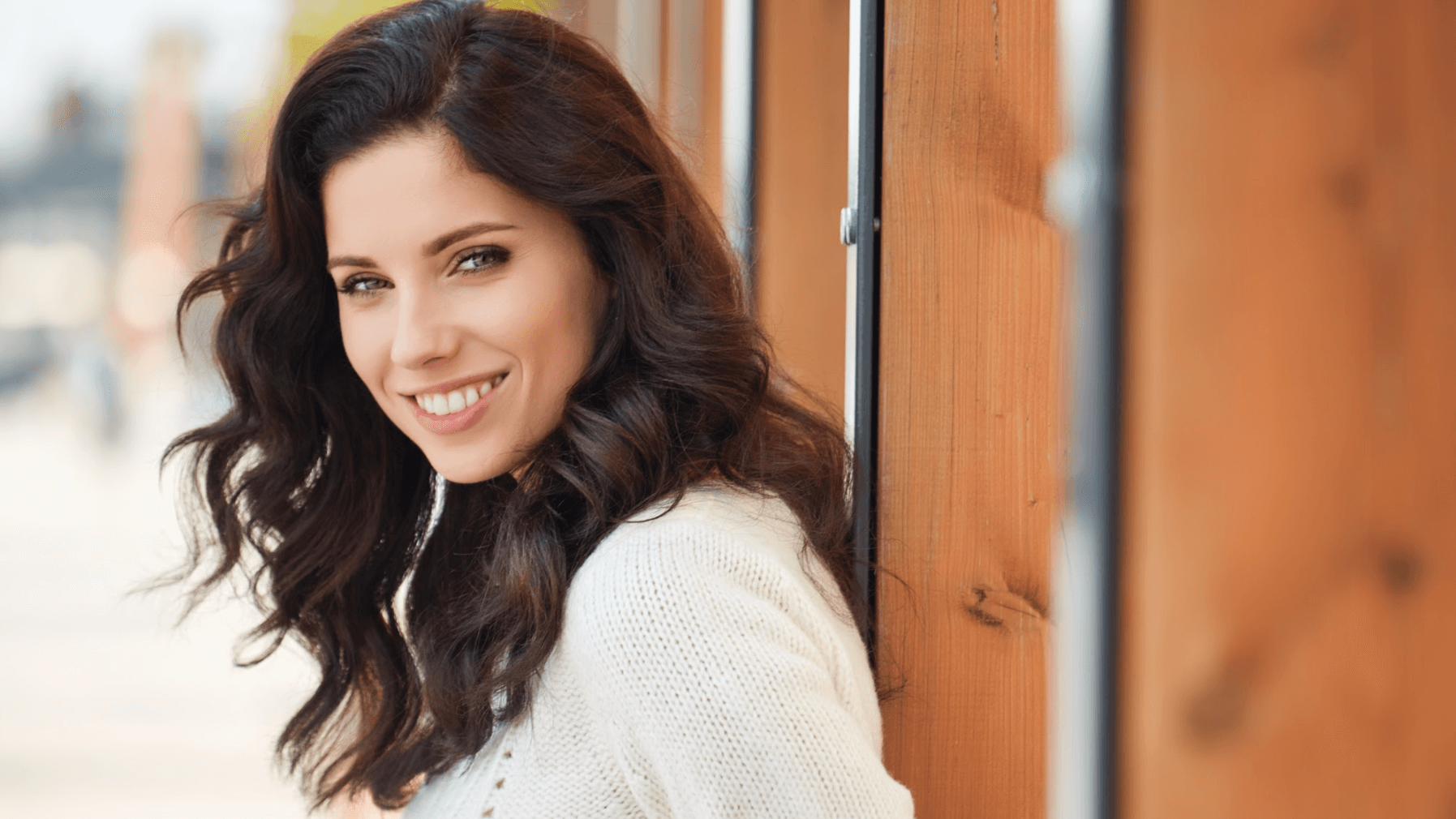 A smiling person with long dark wavy hair stands next to a wooden wall, wearing a textured white top.