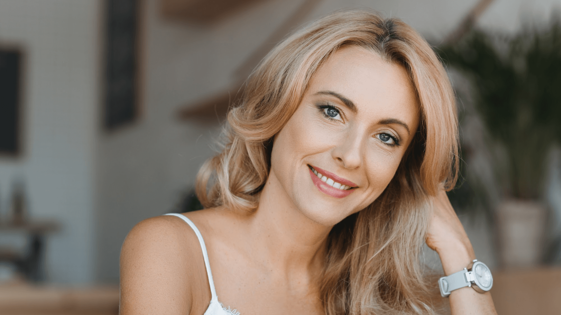 A smiling person with shoulder-length blonde hair, wearing a white top and a watch, posed indoors in a softly lit setting.