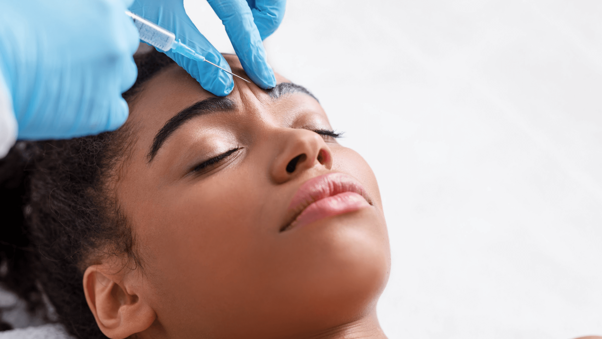 A gloved professional administers a cosmetic injection into the glabellar area between the eyebrows of a person lying down.