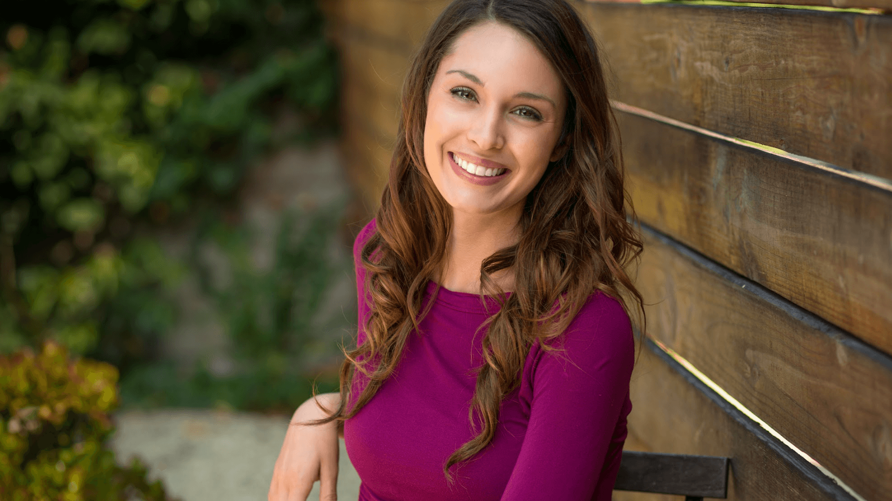 A person with long brown hair, wearing a magenta top, smiles while sitting against a horizontal wooden fence outdoors.
