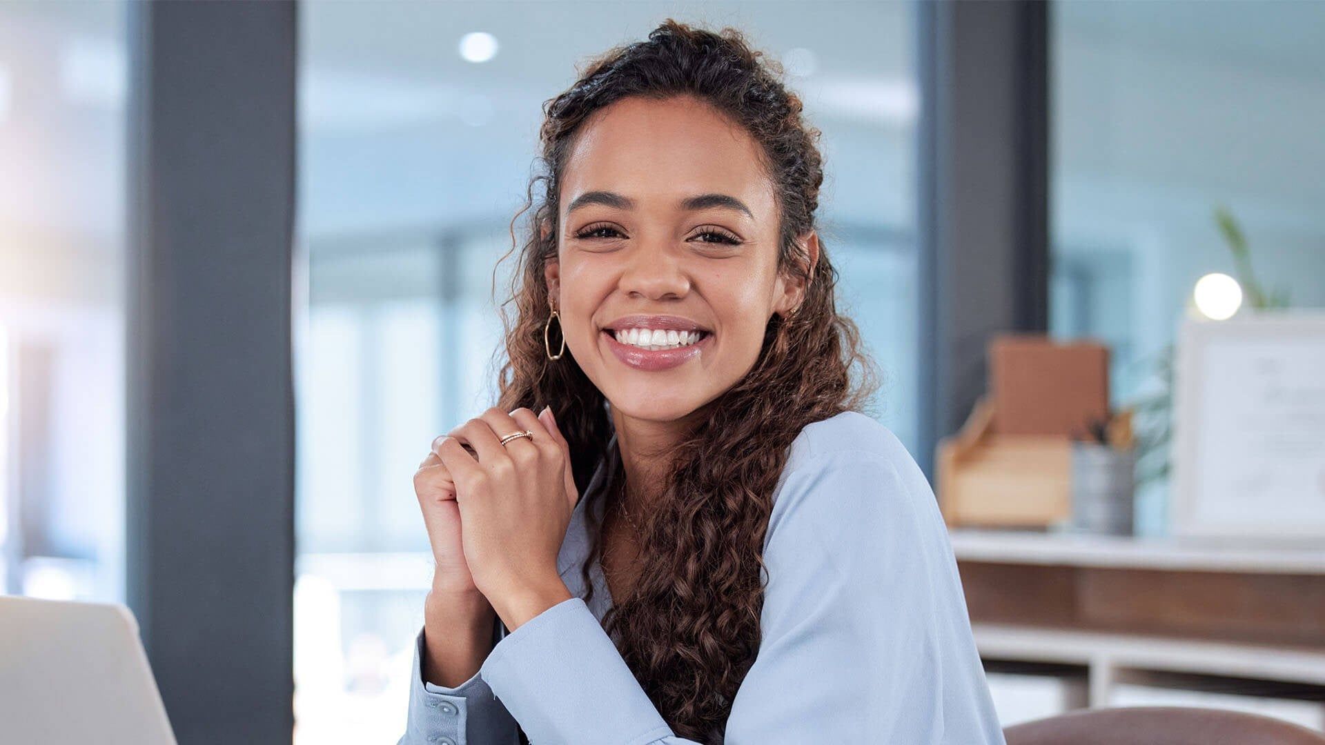 A smiling person with curly hair wears a light blue shirt, sitting with hands clasped in a bright office setting.