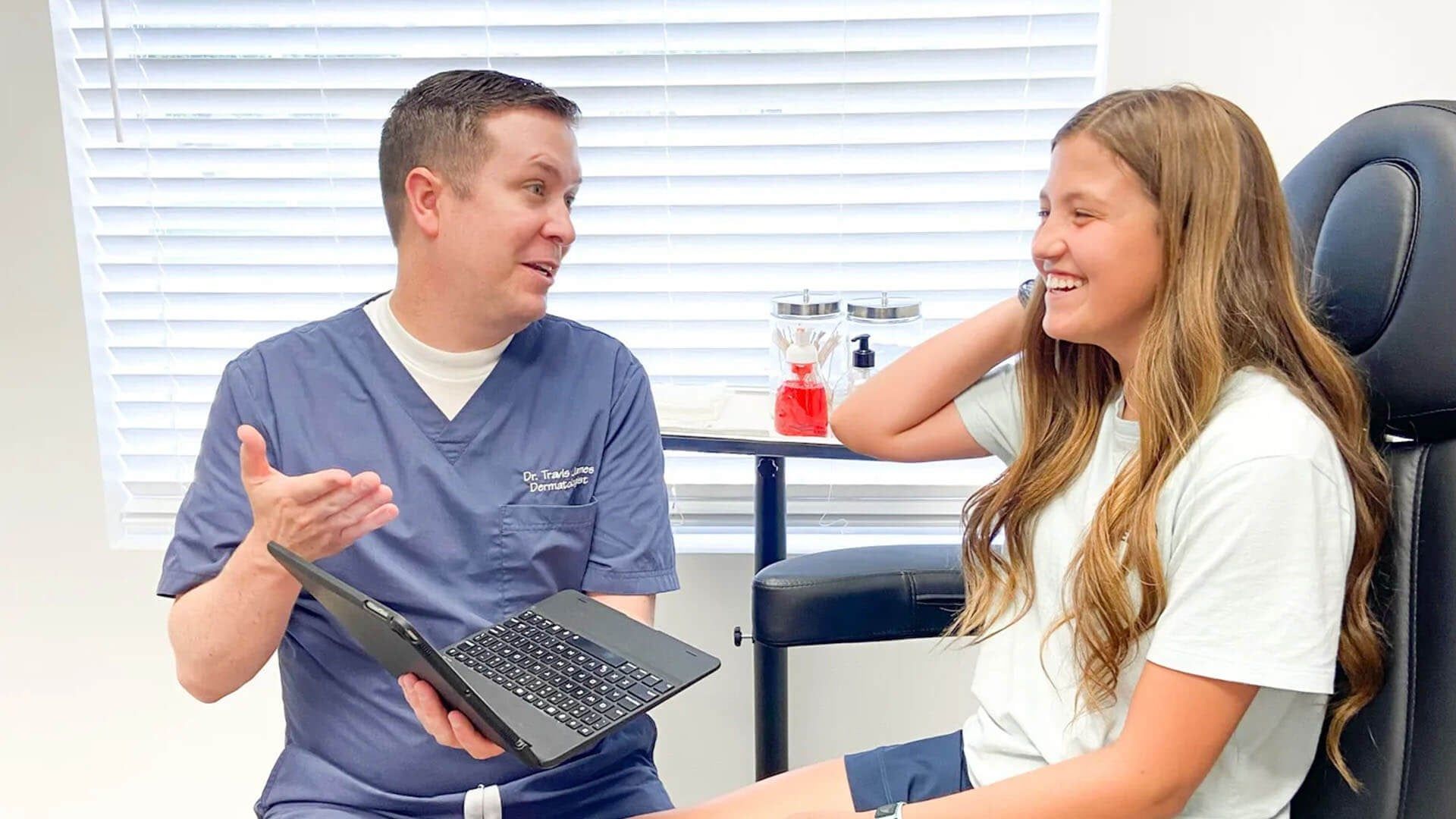 A professional in medical scrubs discusses information on a laptop with a patient in a bright, clinical office setting.