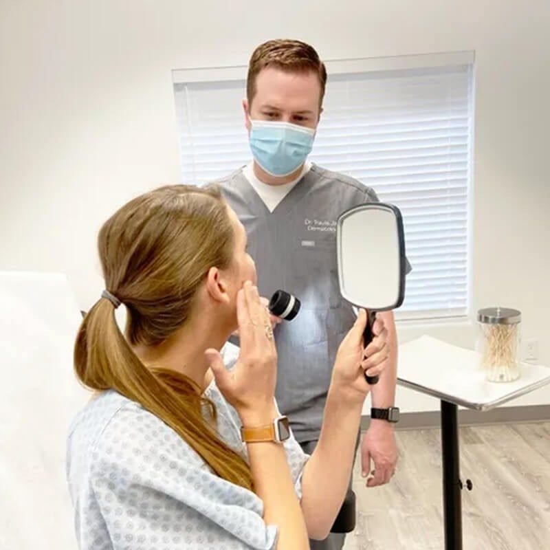 A clinician holding a dermatoscope examines a patient’s face in a bright medical office.