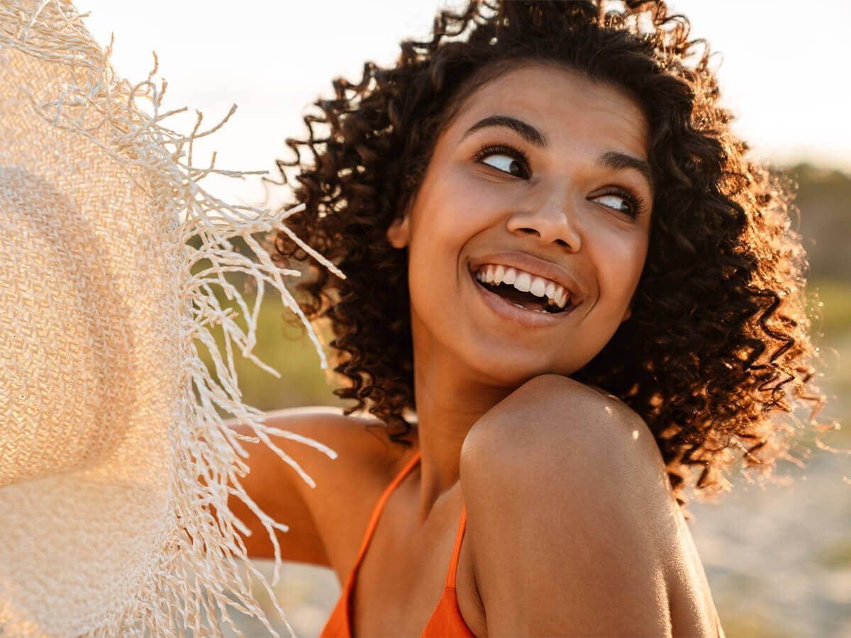 A person with curly hair smiles brightly while holding a wide-brimmed straw hat at the beach during sunset.