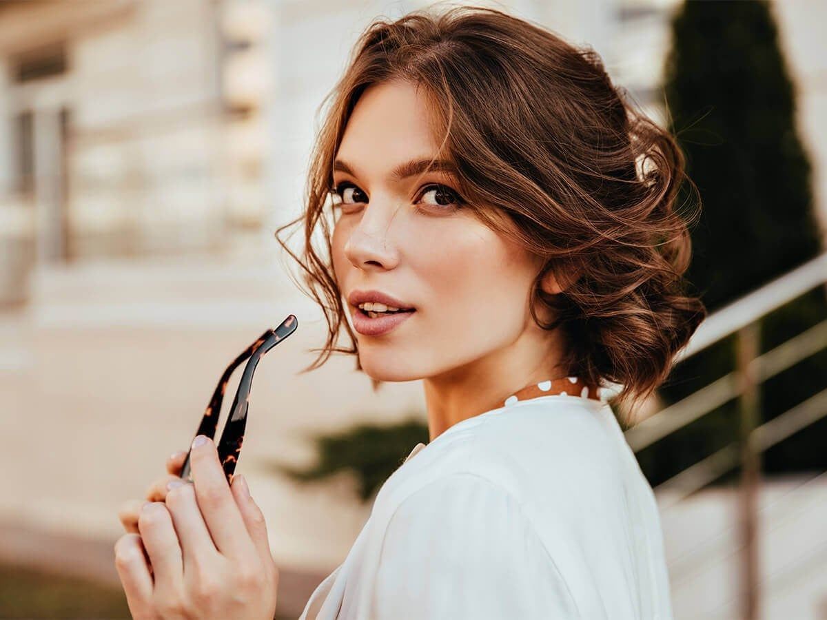 A person with wavy brown hair looks over their shoulder while holding a pair of glasses, set against a blurred background.