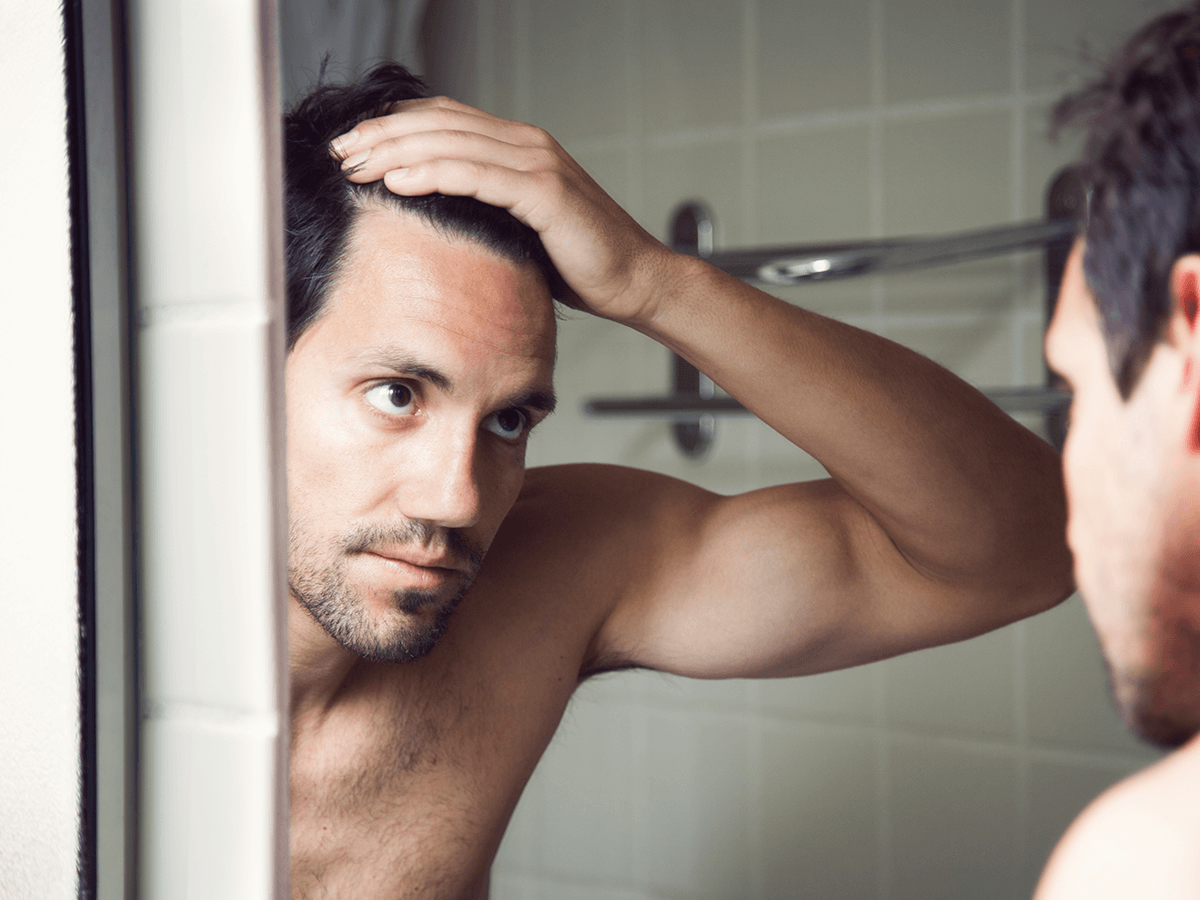 A person looking into a bathroom mirror while pulling back their hair to examine their hairline.