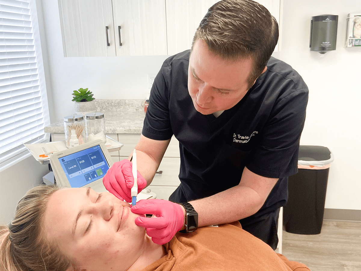 A medical professional in scrubs and pink gloves performs a facial treatment on a patient lying on an exam table.