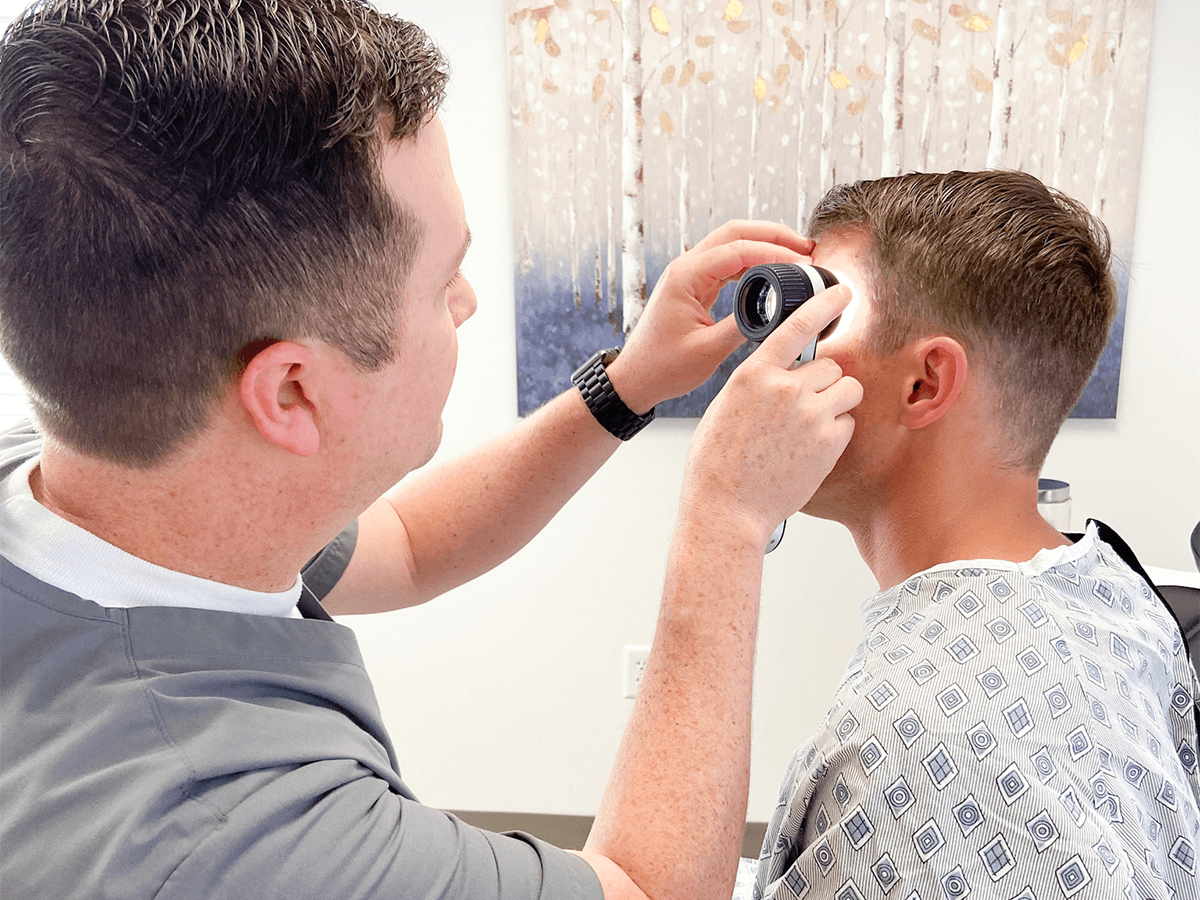 A clinician uses a handheld dermatoscope to examine the skin on a patient's forehead in a medical office.