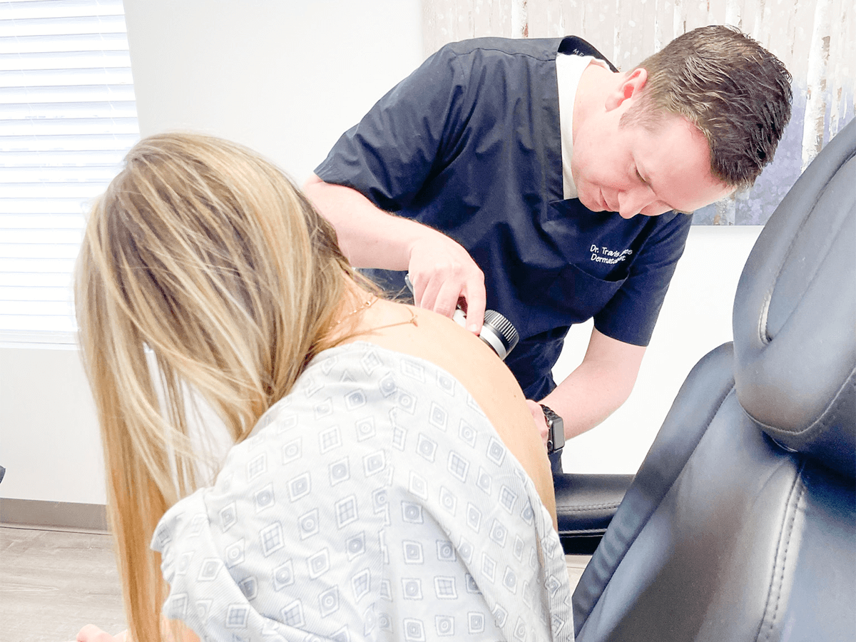 A clinician uses a handheld dermatoscope to examine a spot on a patient's upper back in a medical office setting.