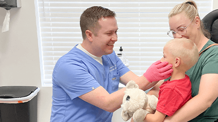 A healthcare worker in blue scrubs wearing pink gloves gently examines a child holding a stuffed bear in a clinic room.
