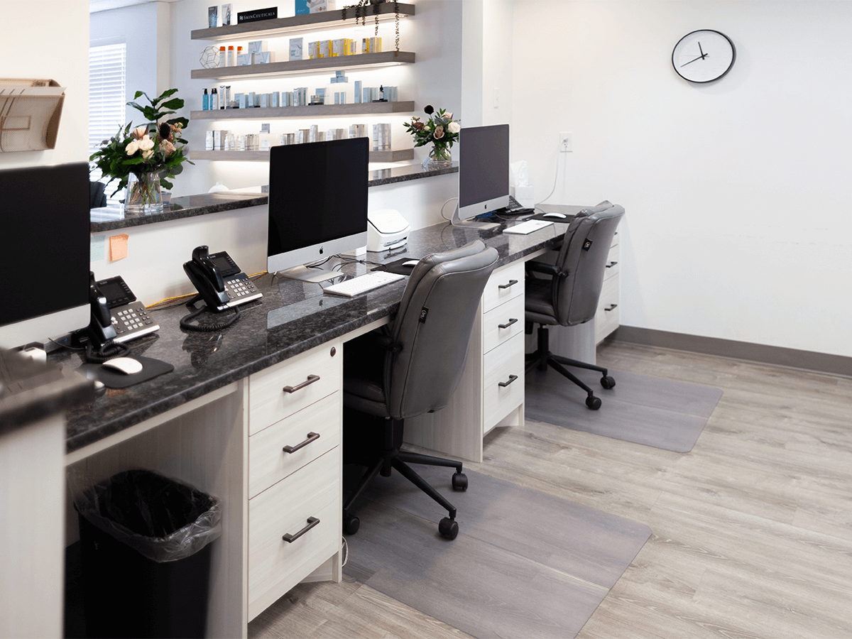 A modern office reception desk with two computer workstations, desk chairs, and product shelves against a white wall.